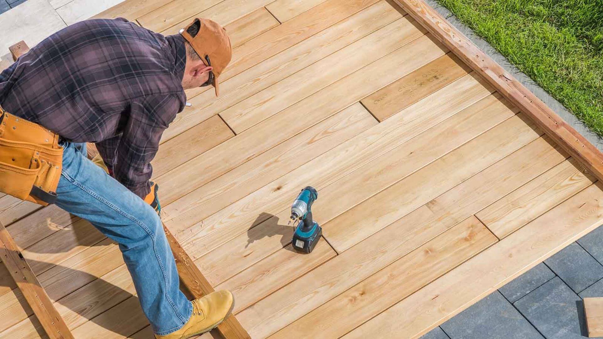 A house is under construction with scaffolding surrounding it. The building is two stories high and features a large, steeply pitched roof. The exterior walls are unfinished, and corrugated metal sheets form a temporary barrier around the site. The foreground shows a grassy area with scattered bushes, and the sky is partly cloudy.